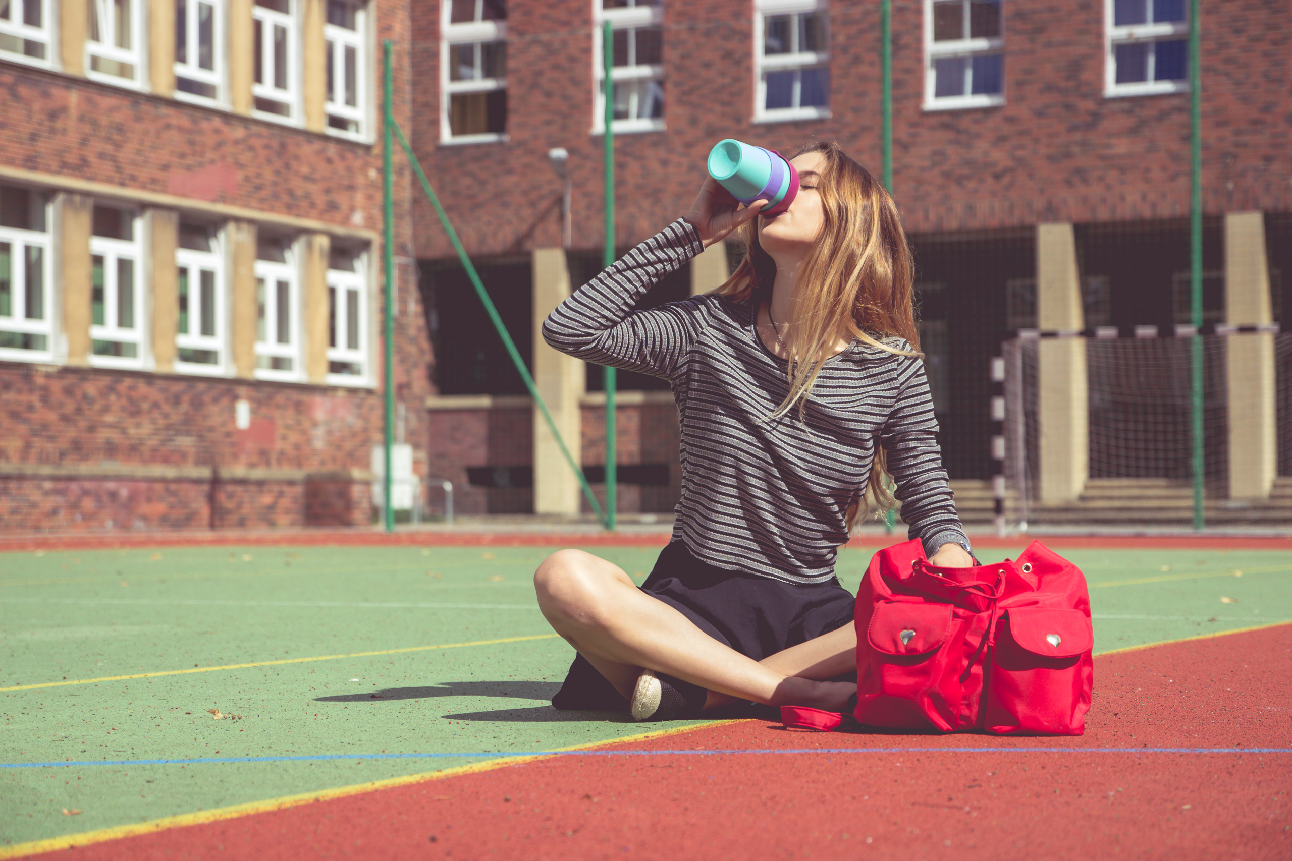 Dziewczyna z czerwonym plecakiem siedzi na boisku szkolnym i piję kawę z niebieskiego kubka