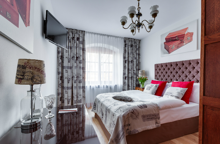 Pokój z dużym podwójnym łóżkiem, utrzymany w kolorystyce czerwono-szarej, duże okno, stolik z lampką