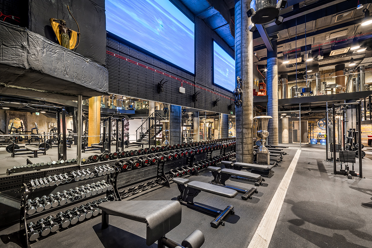 Wnętrze siłowni, dwa duże ekrany oraz przyrządy i urządzenia do ćwiczeń
