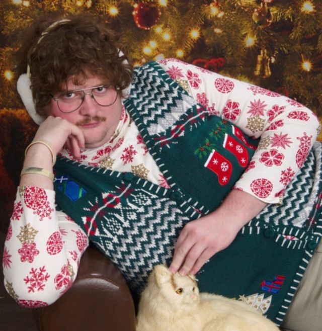 Portret mężczyzny ubranego w kwiecistą koszulę i świąteczny sweter, leżący z kotem na kanapie