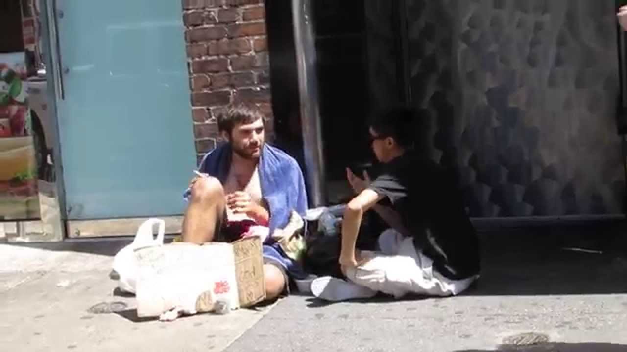 Zdjecie bezdomnego przy ktorym zatrzymal sie mlody czlowiek. Siedza na ulicy i rozmawiaja.