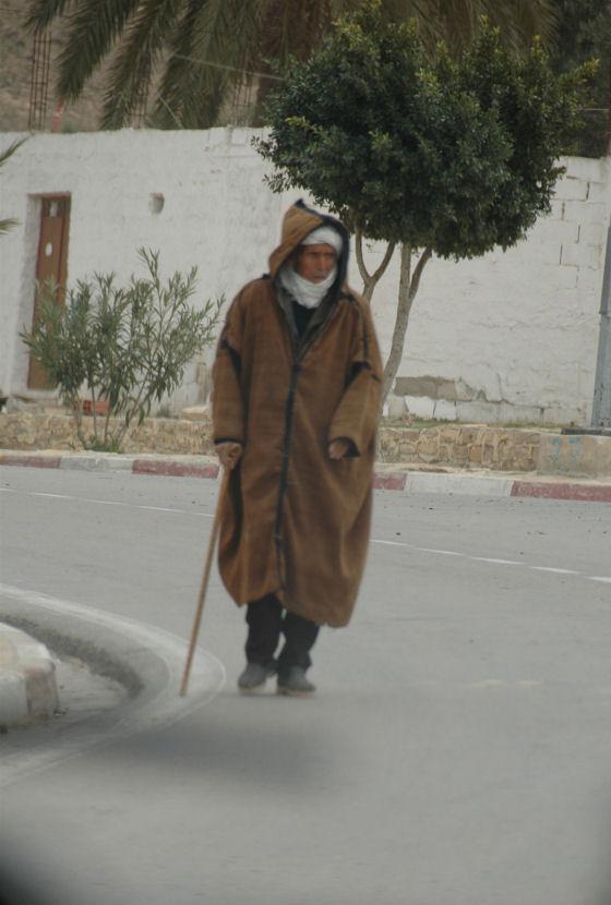 Zdjecie mieszkanca tunezji, ktory idzie ulica. Ubrany jest w zimową galabije, na glowie widac zawiazany turban, w reku trzyma jakąś drewnianą laskę.