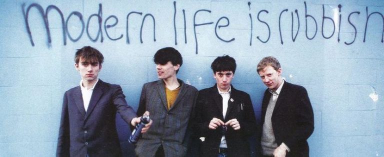 Czterech chłopaków na tle niebieskiej ściany z napisem modern life is robbish