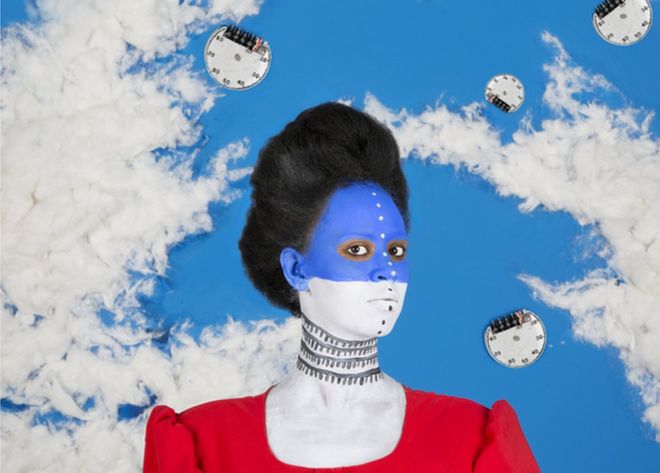 Zdjęcie przedstawia kobietę z półprofilu o pomalowanej na biało błękitnej buzi. W tle widać makietę, która ma przypominać błękitne niebo i chmury