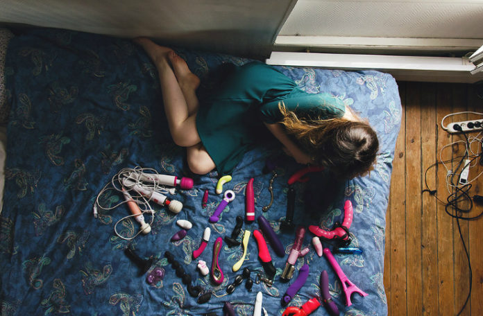 Ujęcie od góry na dziewczynę siedzącą na łóżku, naprzeciwko niej dużo wibratorów