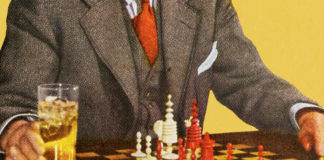 Mężczyzna w graniturze siedzący przy szachach, obok planszy stoi szklanka z napojem