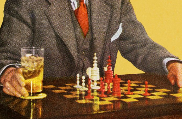 Mężczyzna w graniturze siedzący przy szachach, obok planszy stoi szklanka z napojem