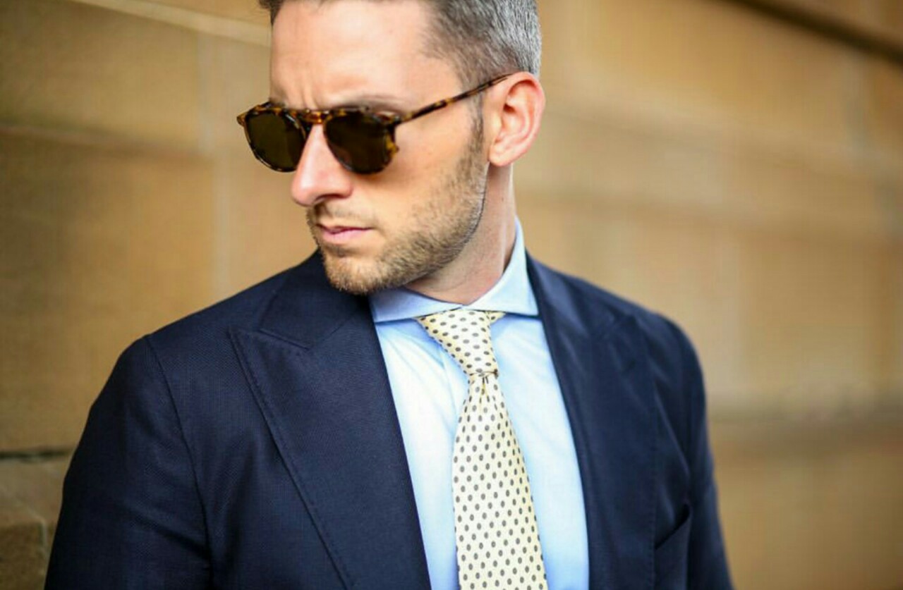 Młody elegancki mężczyzna, w jasnoniebieskiej koszuli, granatowej marynarce z krawatem i w okularach przeciwsłonecznych 