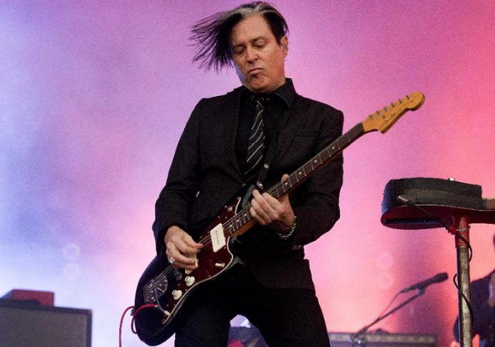 gitarzysta zespołu queens of the stone age, troy van leeuwen podczas koncertu, z gitarą, w czarnej marynarce, czarnej koszuli i czarnym krawacie w drobne białe paski, na różowo-niebieskim tle