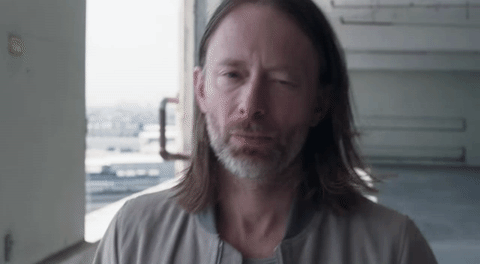 Thom Yorke idący przez garaż, długie przetłuszczone włosy, szara kurtka, delikatny uśmiech