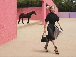 Młoda dziewczyna ubrana w czarną sukienkę, trzyma niewielką torbę. Za nią widać różową ścianę i czarnego konia.