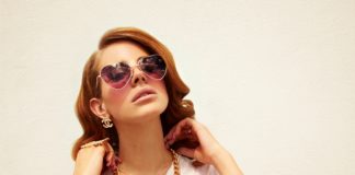 Lana Del Rey w okularach-serduszkach i złotym kajdanie na tle ściany