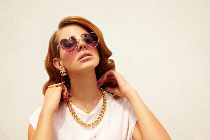 Lana Del Rey w okularach-serduszkach i złotym kajdanie na tle ściany