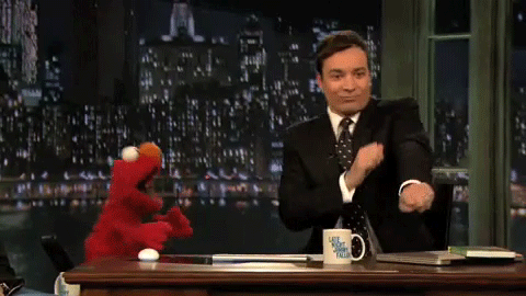 Brunet w garniturze tańczy przy biurku, obok niego czerwony muppet
