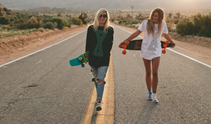 Dwie młode dziewczyny trzymające longboardy, stoją na środku pustej ulicy z deskorolkami
