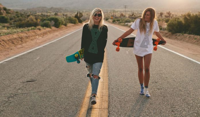 Dwie młode dziewczyny trzymające longboardy, stoją na środku pustej ulicy z deskorolkami