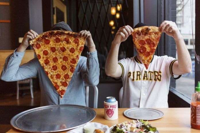 Dwie osoby siedzą przy stoliku i zakrywają sobie twarz wielkimi kawałkami pizzy