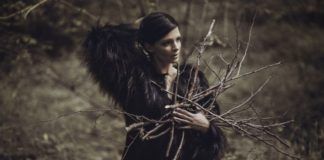 Kobieta z ciemnymi włosami w ciemnym futrze trzyma gałęzie na tle łąki