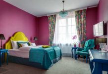 Wnętrze pokoju z ciemno-różowymi ścianami, kolorowym łóżkiem z żółtym oparciem i turkosową narzutą, stolik, telewizor