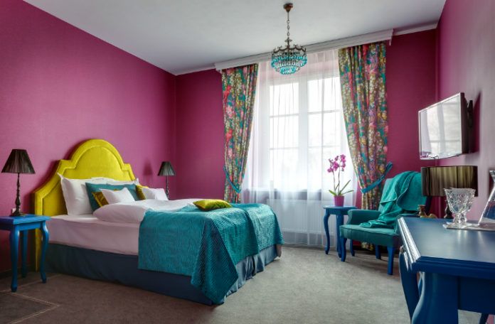 Wnętrze pokoju z ciemno-różowymi ścianami, kolorowym łóżkiem z żółtym oparciem i turkosową narzutą, stolik, telewizor