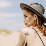 Dziewczyna w szarym kapeluszu ujęta z profilu, patrząca w bok, widoczny tatuaż z różą wiatrów na lewym ramieniu