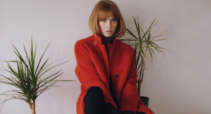 Rudowłosa dziewczyna z grzywką w czerwonym płaszczu siedzaca na szafce