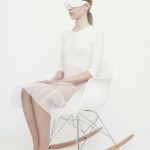 Dziewczyna ubrana na biało, w białych, zakrywających całkowicie oczy okularach, siedzi na białym krześle z drewnianymi elementami