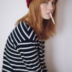 Dziewczyna z grzywką w czerwonej czapce i bluzce w paski siedząca profilem z papierosem w ustach