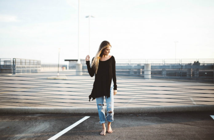 Dziewczyna stojąca boso na molo ubrana w poszarpane jeansy i czarną bluzkę