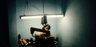 Zdjęcie kolorowe utrzymane w ciemnych barwach. Na środku, wśród przyrządów do obróbki tkanin siedzi kobieta. Zza maszyny ledwo widać połowę jej twarzy, drugą połowę przysłania ciemna grzywka.