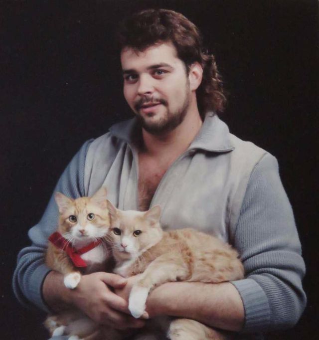 Mężczyzna w jeansowej lekko rozpiętej koszuli trzyma na rękach dwa koty