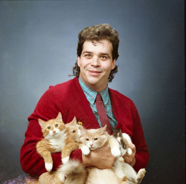 Portret mężczyzny w czerwonym swetrze, koszuli i krawacie, trzymający cztery koty