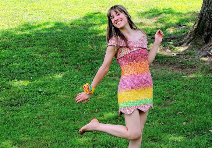 Na zdjęciu widzimy artystkę, która prezentuje sukienkę wykonaną z opakowań po cukierkach. Stoi na tle trawy i uśmiecha się.