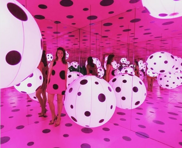 Zdjęcie przedstawia artystkę na tle różowego pomieszczenia, w którym są duże białe kule w duże czarne kropki. Artystka ma na sobie sukienkę w tym samym wzorze.