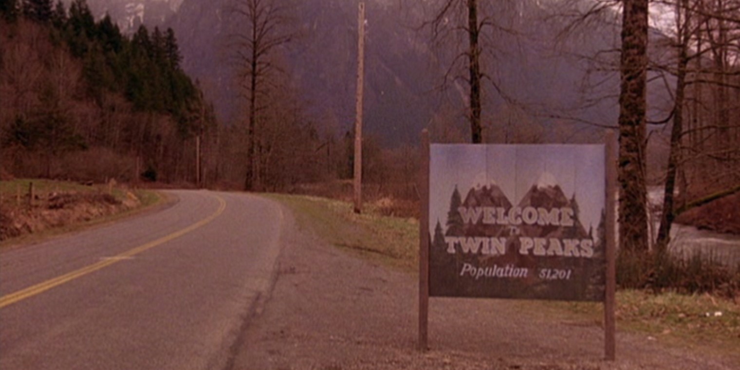 Wjazd do miasteczka na tablicy widzimy napis Welcome To Twin Peaks Po lewej stronie znajduje sie ulica a po prawej drzewa i zielona trawa