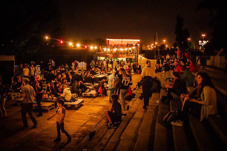 Zdjęcie przedstawia bulwar nad wisłą wieczorem. Ludzie siedzą na schodkach przy rzece, w oddali widać scenę klubu "Cud nad Wisłą".