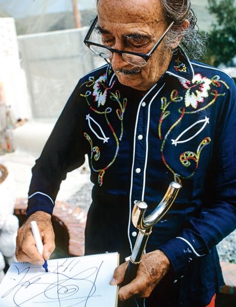 Na zdjęciu widzimy Salvadora Dalego, który ma okulary, i stoi szkicując.