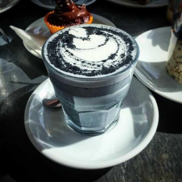 Na zdjęciu jest kubek z kawą latte ciemnego koloru, zaś na nim artystycznie spienione mleko. Kubek stoi na białym talerzyku, a obok są inne talerzyki.