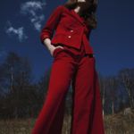Dziewczyna stojąca na trawie, w tle las, ubrana w czerwone spodnie i czewoną marynarkę