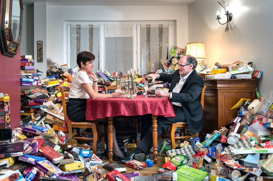 zdjęcie przedstawia małżeństwo siedzące przy stole, a wokół nich jest cały pokój zagracony pustymi opakowaniami po produktach codziennego użytku