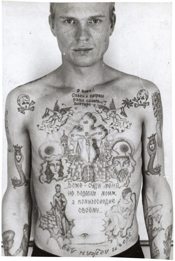 Zdjęcie męzczyzny widocznego od pasa w górę. Jego cała klatka piersiowa oraz ramiona są wytatuowane w radzieckie, więzienne wzory. Są motywy religijne, napisy cyrylicą oraz czaszki. Męzczyzna stoi na tle białej ściany.