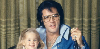 Zdjecie Elvisa Presley, ktory siedzi na krzesle ubrany w niebieski dres, w okularach, trzyma mikrofon w jednym reku, a drugą ręką trzyma siedzącą na kolanach córkę. Dziewczynka jest jeszcze w wieku przedszkolnym. W przyszlosci zostanie zona Micheala Jacksona.
