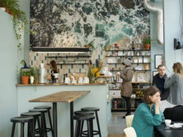 Zdjęcie przedstawia kawiarnię w środku, ladę, baristkę oraz osoby siedzące w kawiarni