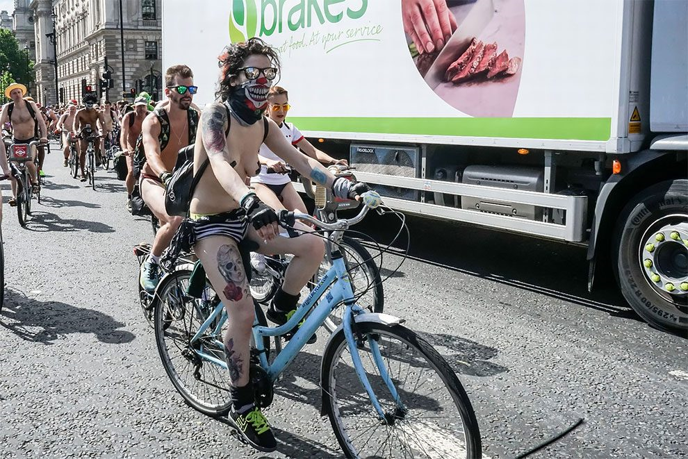 Na zdjęciu widać wytatuowana, białą dziewczynę, która jedzie naga na rowerze w ramach World Naked Bike Ride. Za nią widać kolejnych uczestników oraz stojący na poboczu samochod dostawczy.