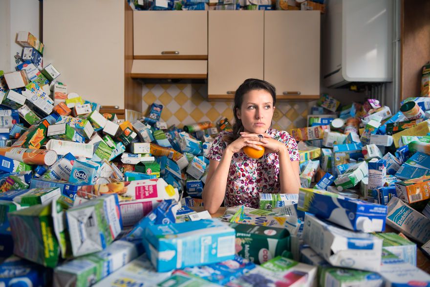 zdjęcie przedstawia kobietę siedzącą w kuchni a wokół niej jest pełno pustych opakowań po mleku zajmujących całą kuchnię