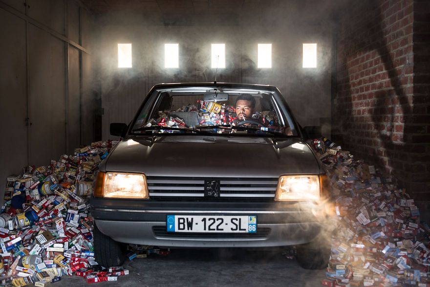 zdjęcie przedstawia mężczyznę siedzącego w samochodzie w garażu, a wokół samochodu jest pełno pustych opakowań po papierosach