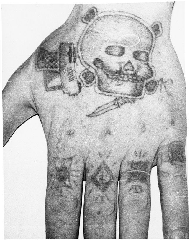 Czarno-białe zdjęcie nadgarstka, na którym są więzienne tatuaże: czaszka, pistolet, nóż, litera k, oraz wydziarane pierścienie na palcach.