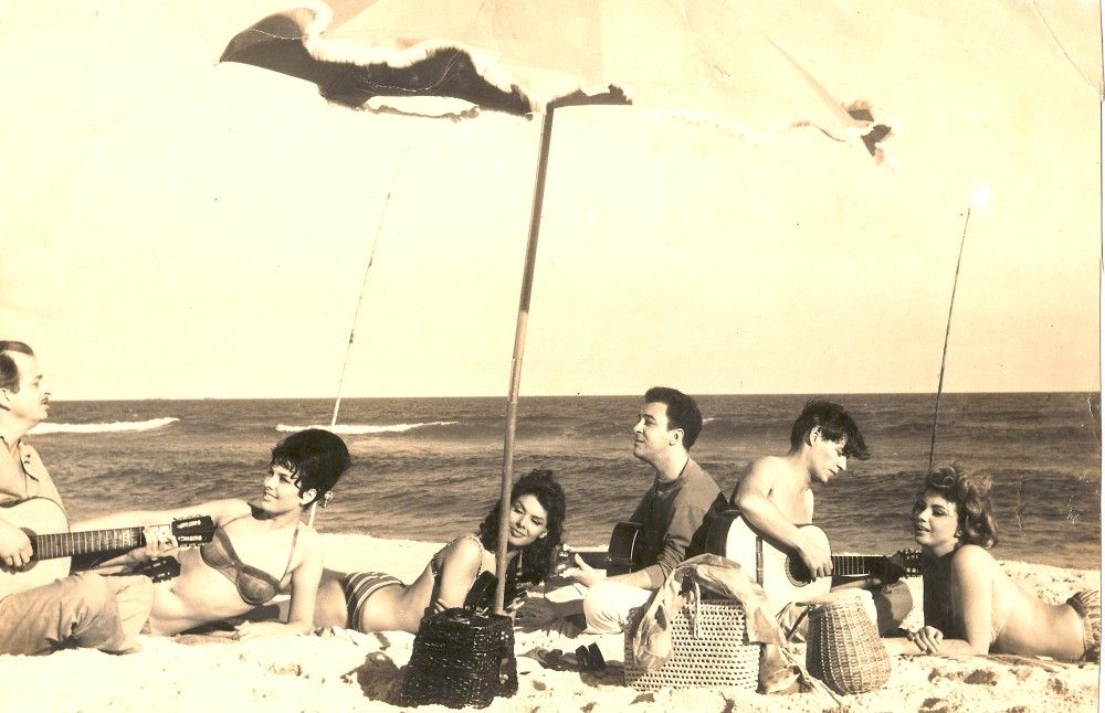 Zdjęcie w stylu sepia. Widoczna jest grupka znajomych, trzech mezczyzn i trzech kobiet lezacych pod parasolem na plazy. Trzej mezczyzni zabawiaja damy grając muzykę na gitarach i śpiewając.