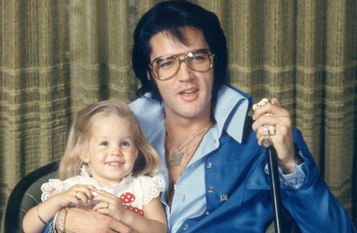 Zdjecie Elvisa Presley, ktory siedzi na krzesle ubrany w niebieski dres, w okularach, trzyma mikrofon w jednym reku, a drugą ręką trzyma siedzącą na kolanach córkę. Dziewczynka jest jeszcze w wieku przedszkolnym. W przyszlosci zostanie zona Micheala Jacksona.