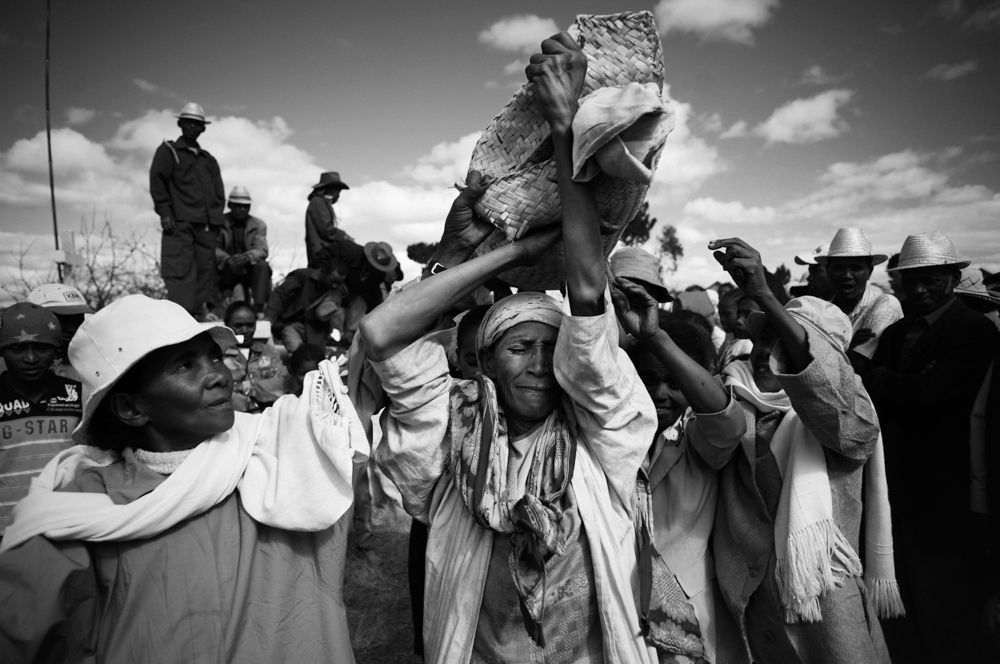 Zdjęcie ludu mieszkajacego na Madagaskarze, który kontynuuje tradycję famadihana. Wyjmują zmarłego z grobu, owijają nowymi całunami i chowają jeszcze raz. Na zdjęciu widać bliskich nieboszczyka, który trzymają go na rękach owiniętego w biały koc.